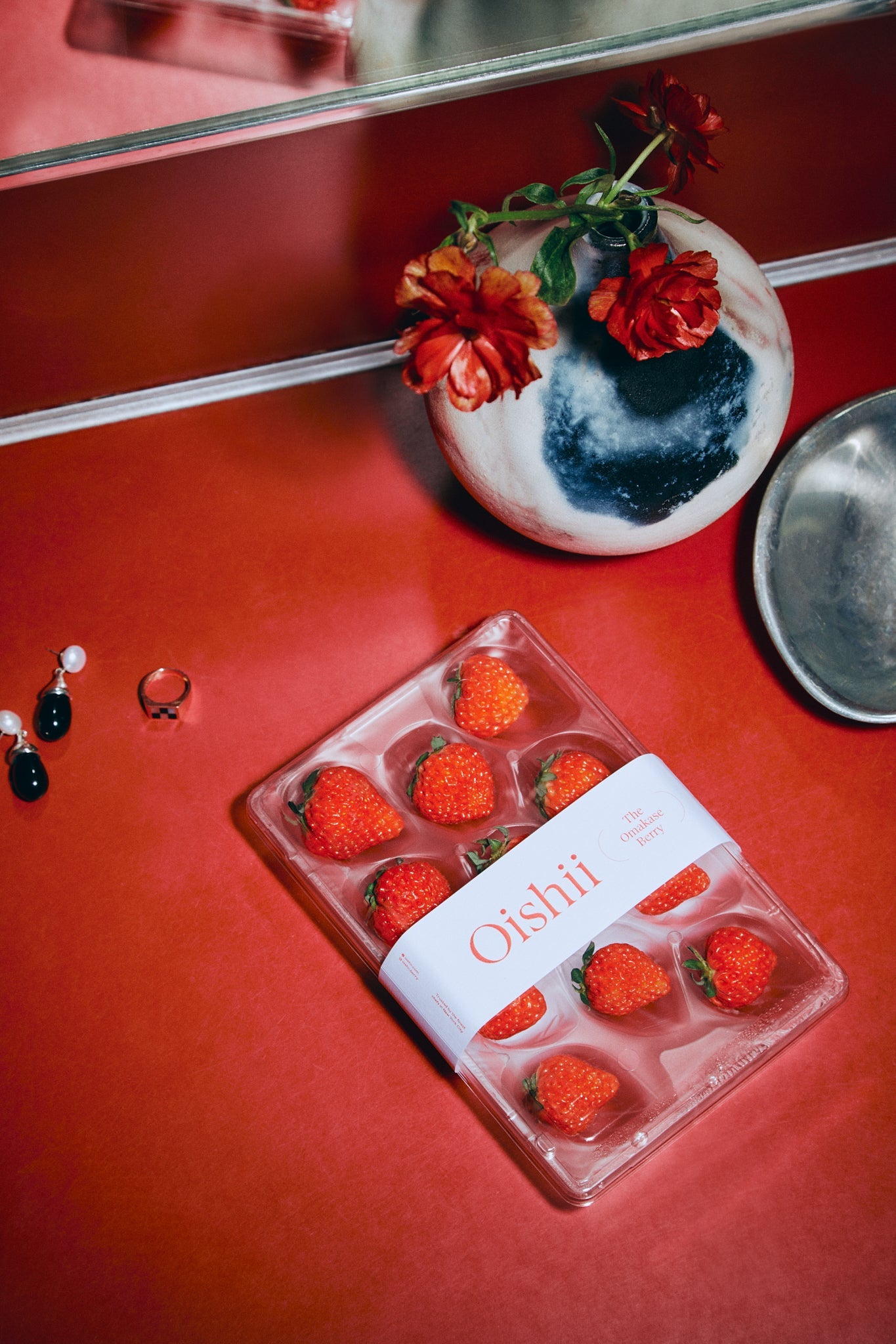 An 11 berry tray of Omakase Berries lays on a red countertop next to a pair of black and white earrings, a ring, a silver dish, and two red flowers in a round, white and blue vase.